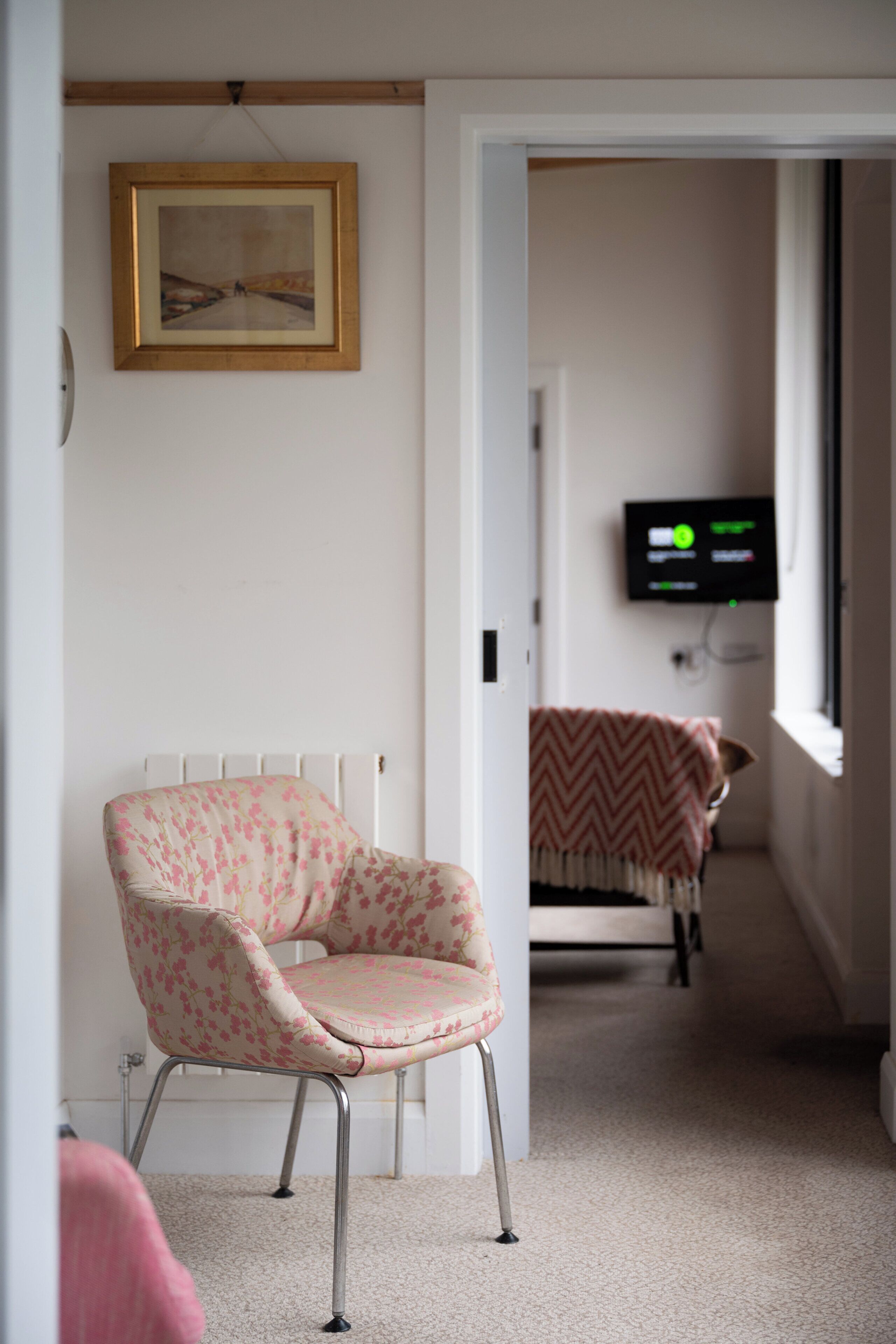 A room with a floral-patterned chair, a framed picture on the wall, a television on the wall, and a bed with a chevron-patterned blanket.
