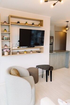 A living room with a flat-screen TV, a sofa, a small table, and a kitchen area in the background. - Newly Renovated 2BR Apartment (Lince)