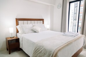 A neatly made bed with a wooden headboard, a bedside lamp, and a view of a cityscape through a window. - Newly Renovated 2BR Apartment (Lince)