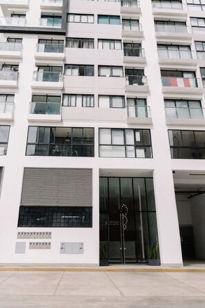 A modern multi-story building with a flat facade, large windows, and a ground-level entrance with a glass door. - Newly Renovated 2BR Apartment (Lince)