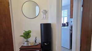 A room with a round mirror, a black water filter dispenser, a small table with a potted plant, a candle, and a sign indicating free filtered water. - Modern 2BR • Steps to Jean-Talon Metro • Fast Wi-Fi • Long Stays Welcome (Montréal)