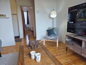 Ein Wohnzimmer mit Fernseher, auf dem eine Sendung läuft, einem Sessel mit Kissen, einer Bärenfigur und zwei Tassen auf dem Boden.