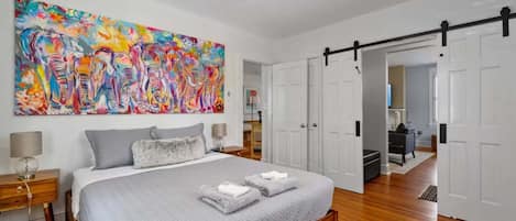 Un dormitorio con una cama grande, un ventilador de techo, un cuadro abstracto colorido y una puerta corredera que da a otra habitación.