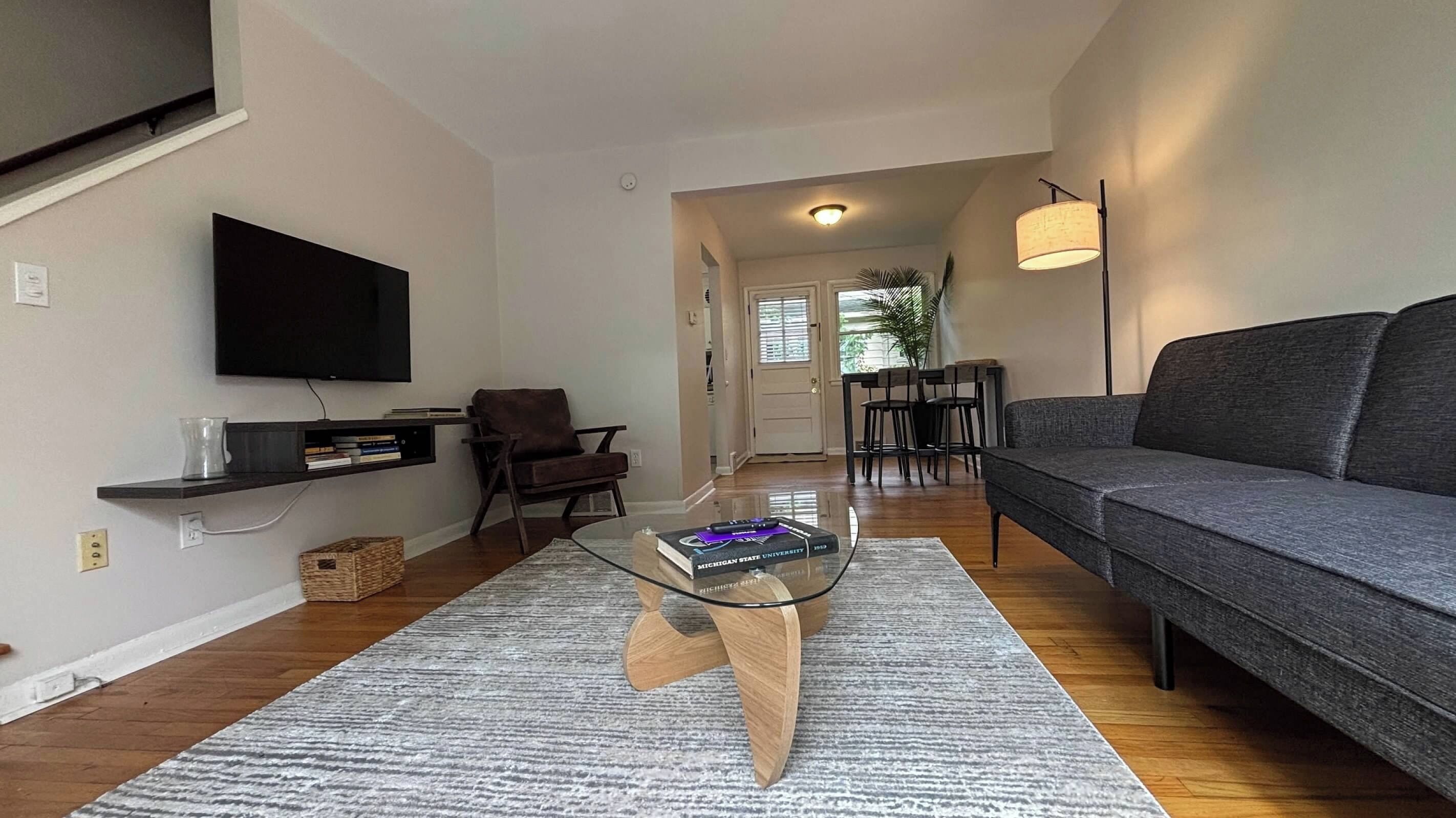 A modern living room with a flat-screen TV, a dark grey sofa, a glass coffee table, and a dining area with chairs.