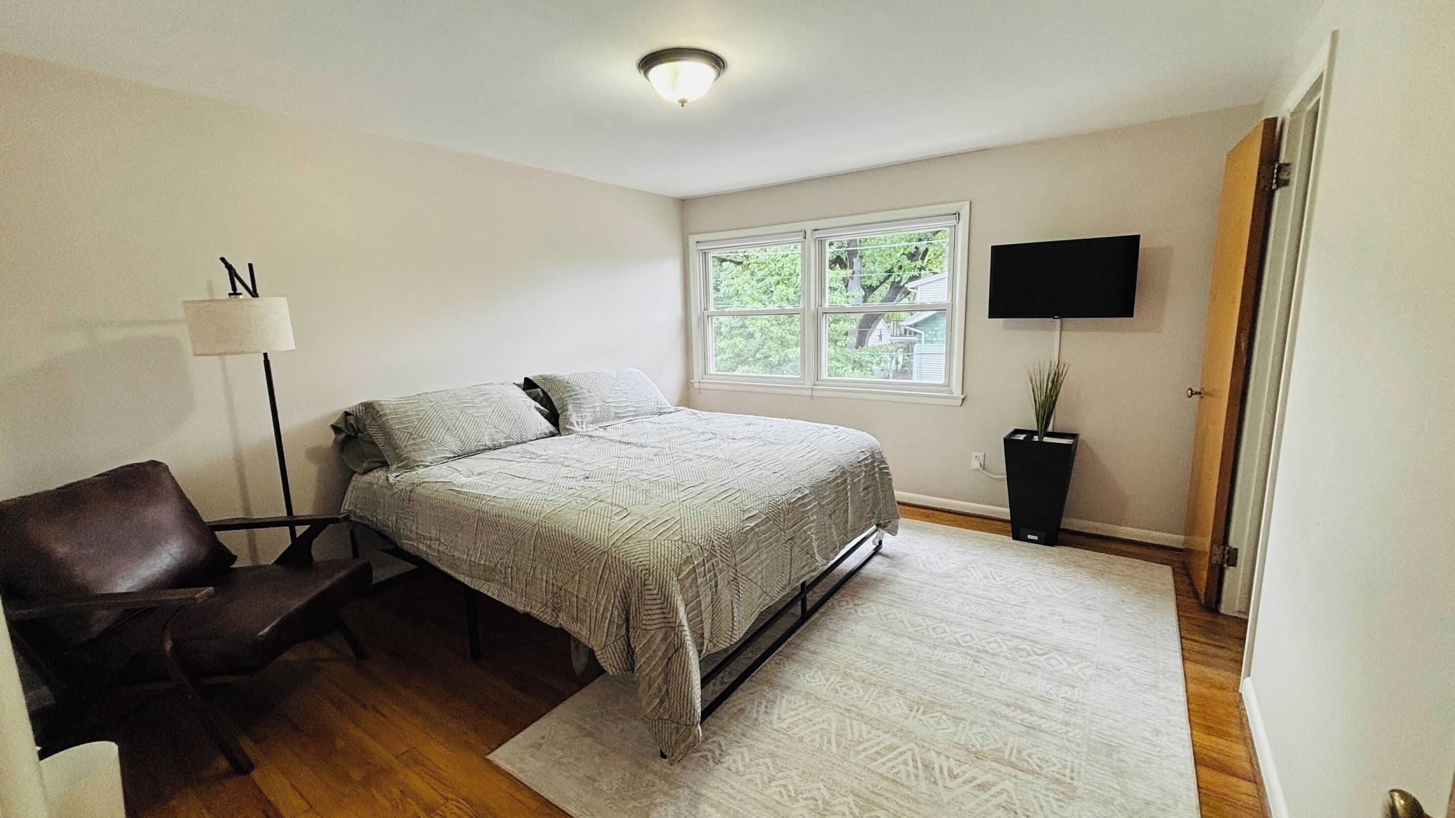 A bedroom with a bed, a desk, a chair, a floor lamp, a TV, and a window with a view of greenery.