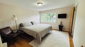 A bedroom with a bed, a desk, a chair, a floor lamp, a TV, and a window with a view of greenery.