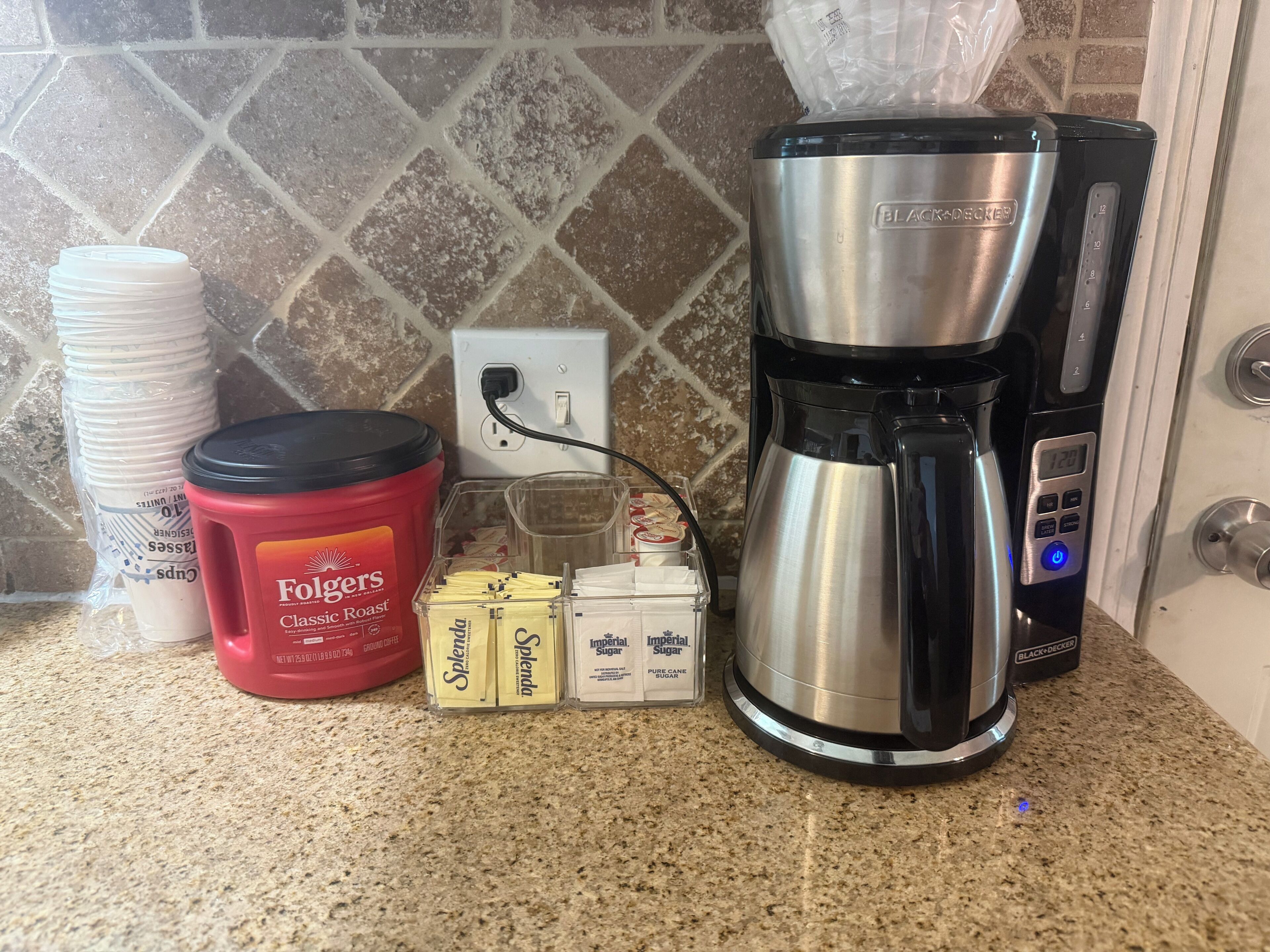 Un comptoir de cuisine avec une machine à café, un récipient rouge étiqueté « Folgers Classic Roast », une pile de gobelets en papier blancs et des boîtes de Splenda et de sucre Imperial.