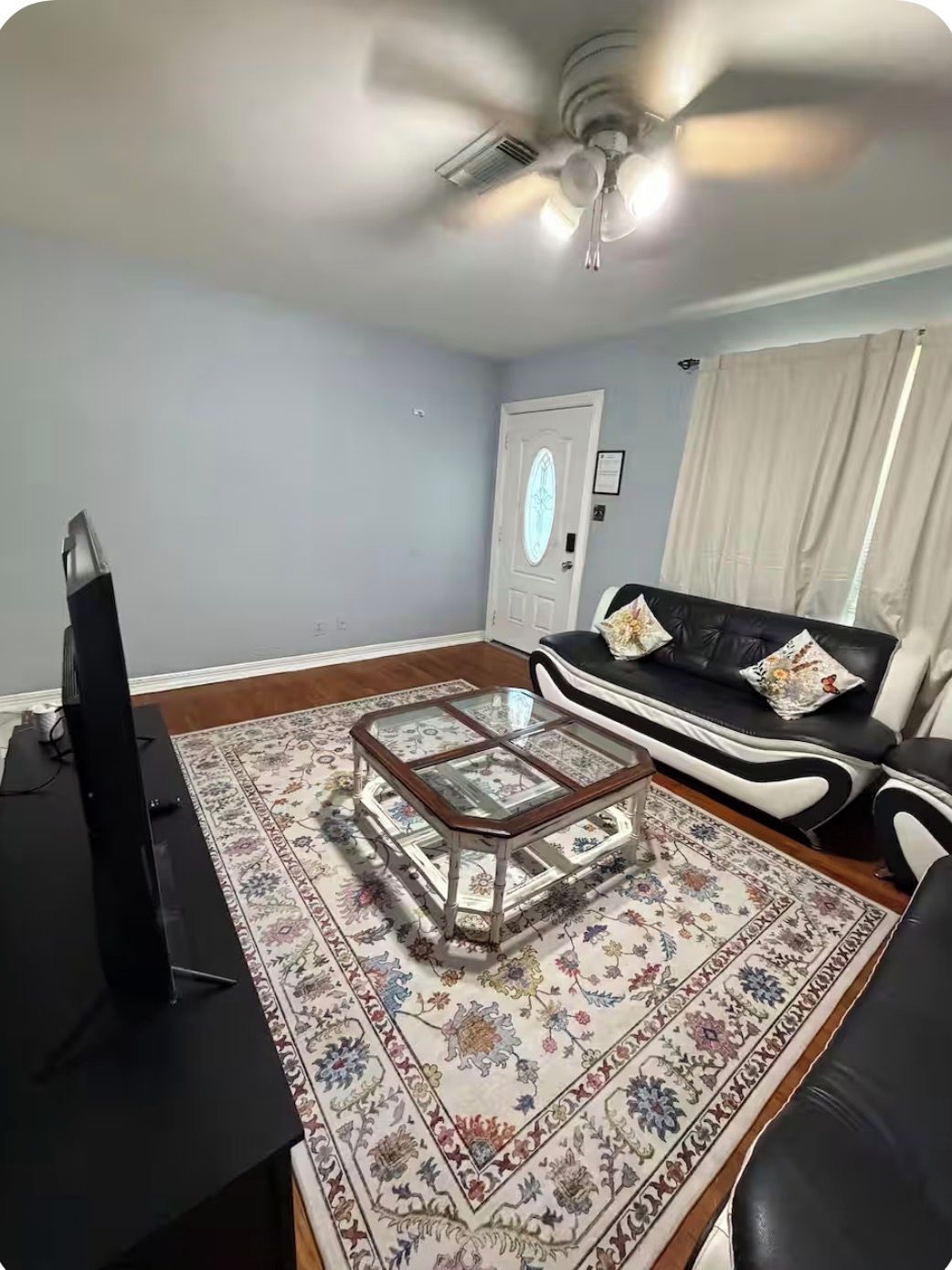 A living room with a television, a glass coffee table, a black sofa, and a patterned rug.