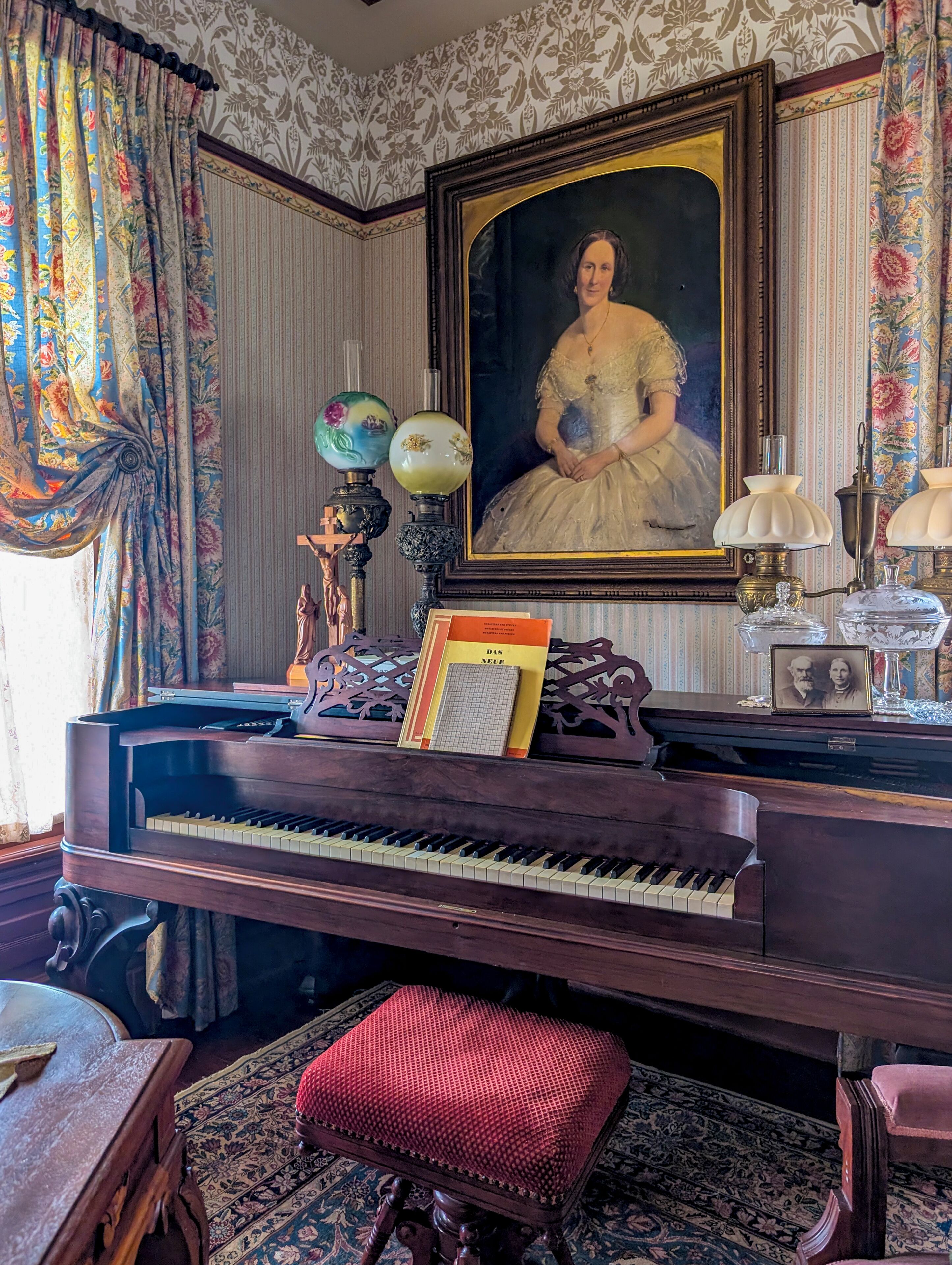 Una habitación con un piano de cola, un retrato enmarcado, un globo terráqueo, una lámpara y un taburete tapizado de rojo.