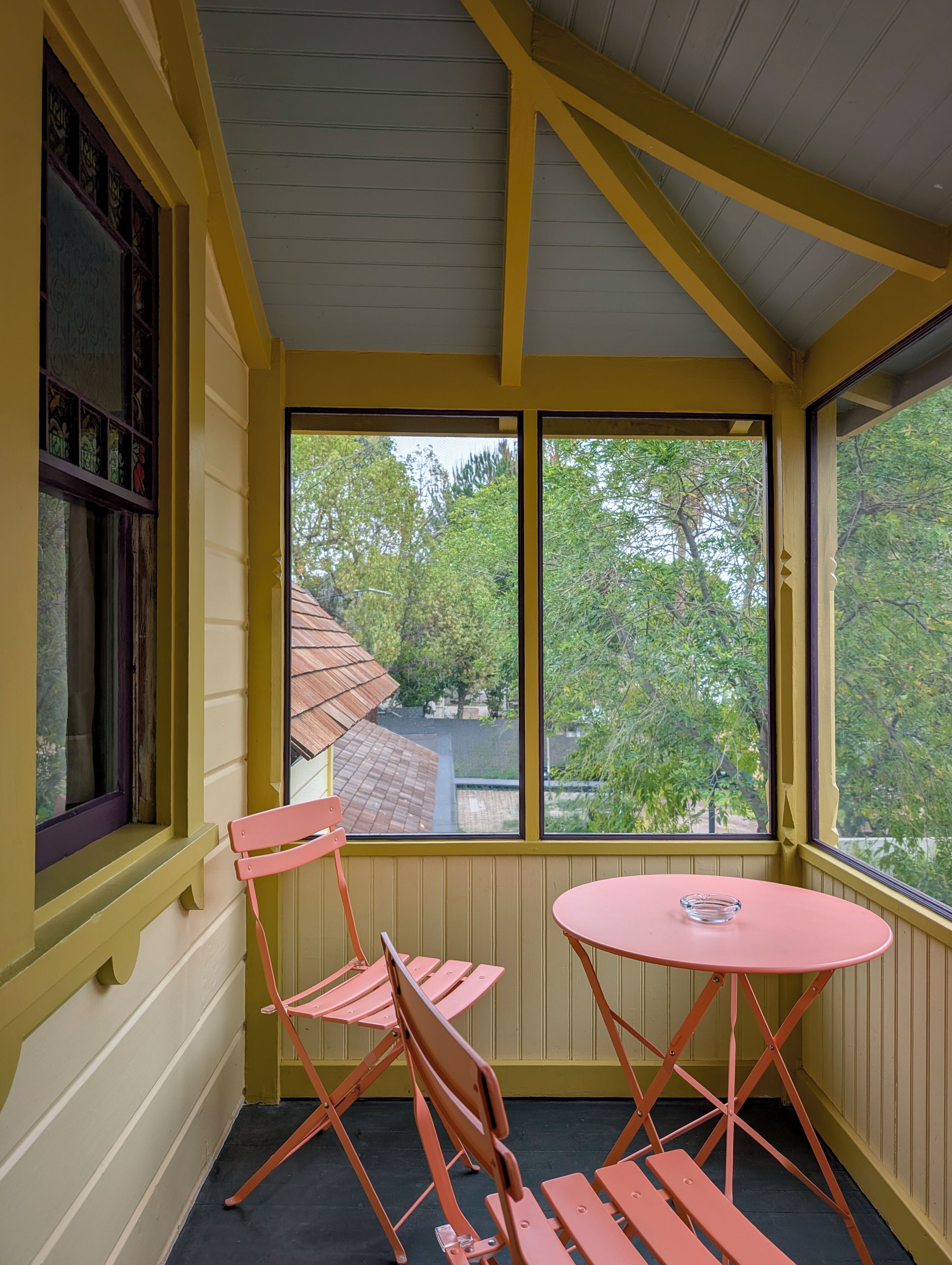 Una habitación con paredes amarillas, una ventana con vista a árboles y una mesa rosa con un vaso.