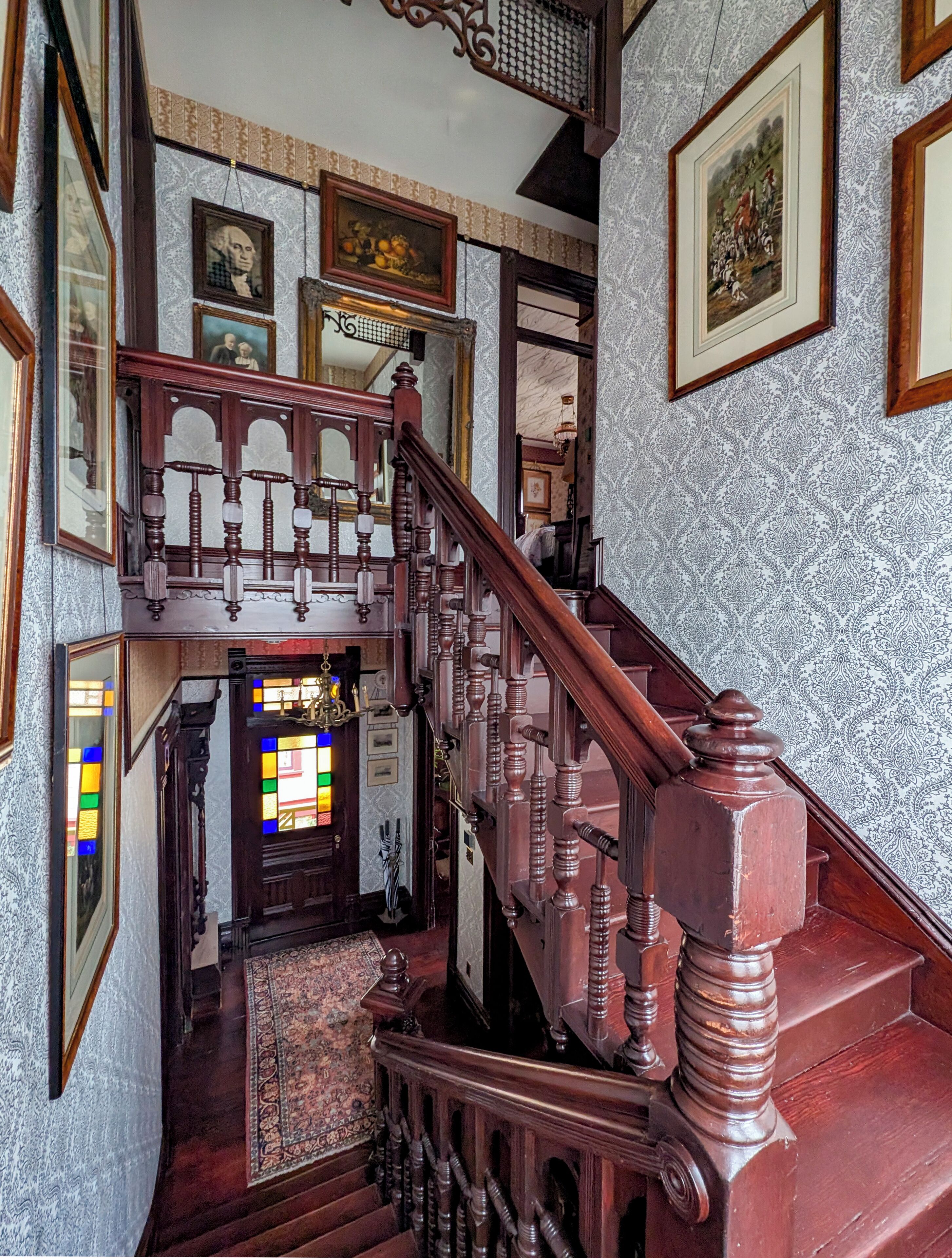 Una imponente escalera con ornamentada carpintería, que conduce a un espacio con un vitral y obras de arte enmarcadas en las paredes.