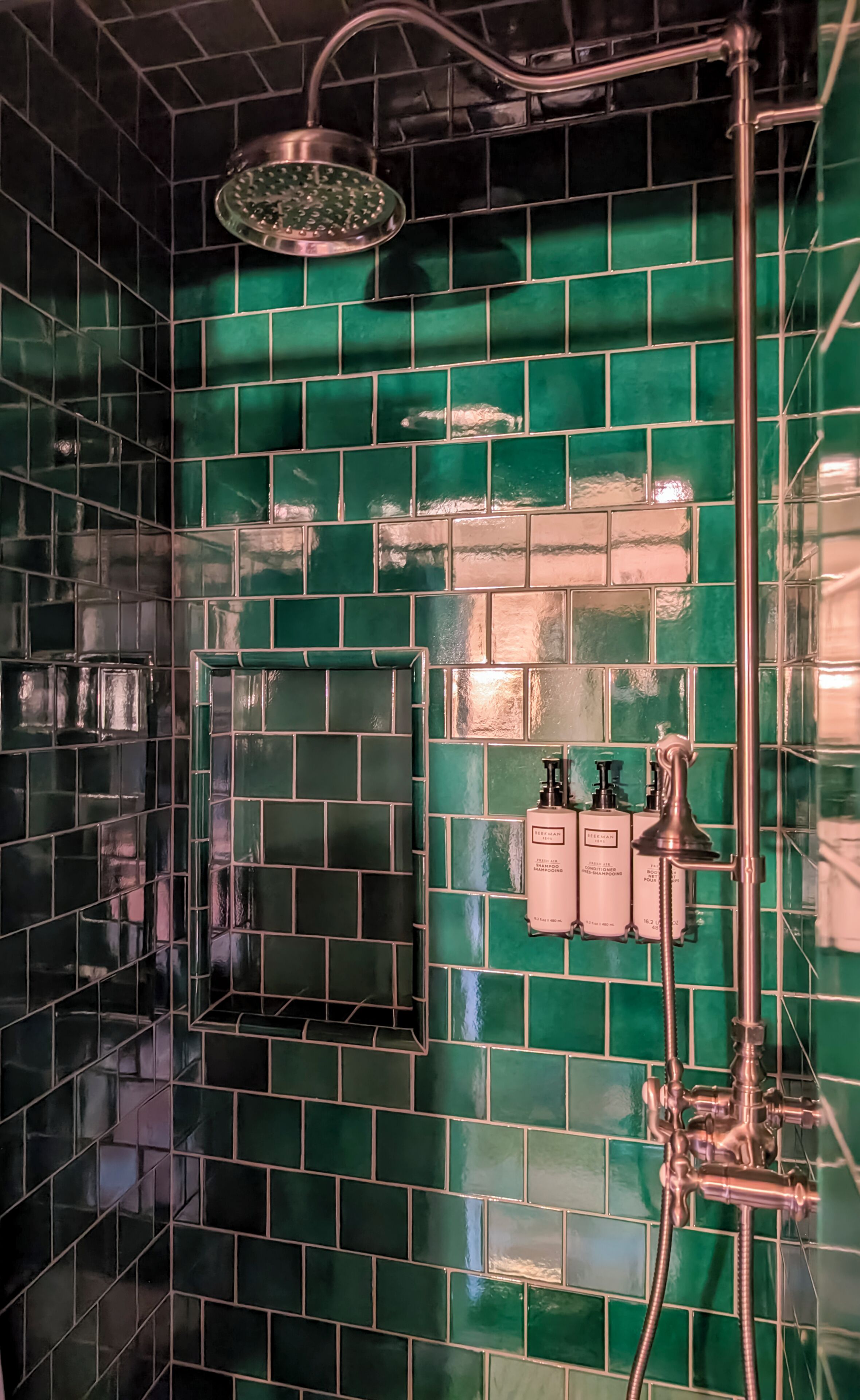 A bathroom with green mosaic tiles, a showerhead, and two shampoo dispensers.