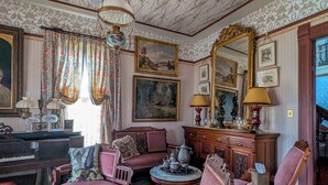 A room with a piano, multiple chairs, a round table with a teapot, and a large mirror.