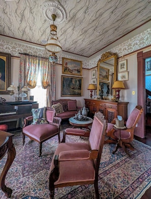 A room with a piano, multiple chairs, a round table with a teapot, and a large mirror. - The Arthur Blair Historic Inn (Monrovia)