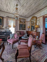 Una habitación con un piano, varias sillas, una mesa redonda con una tetera y un espejo grande.