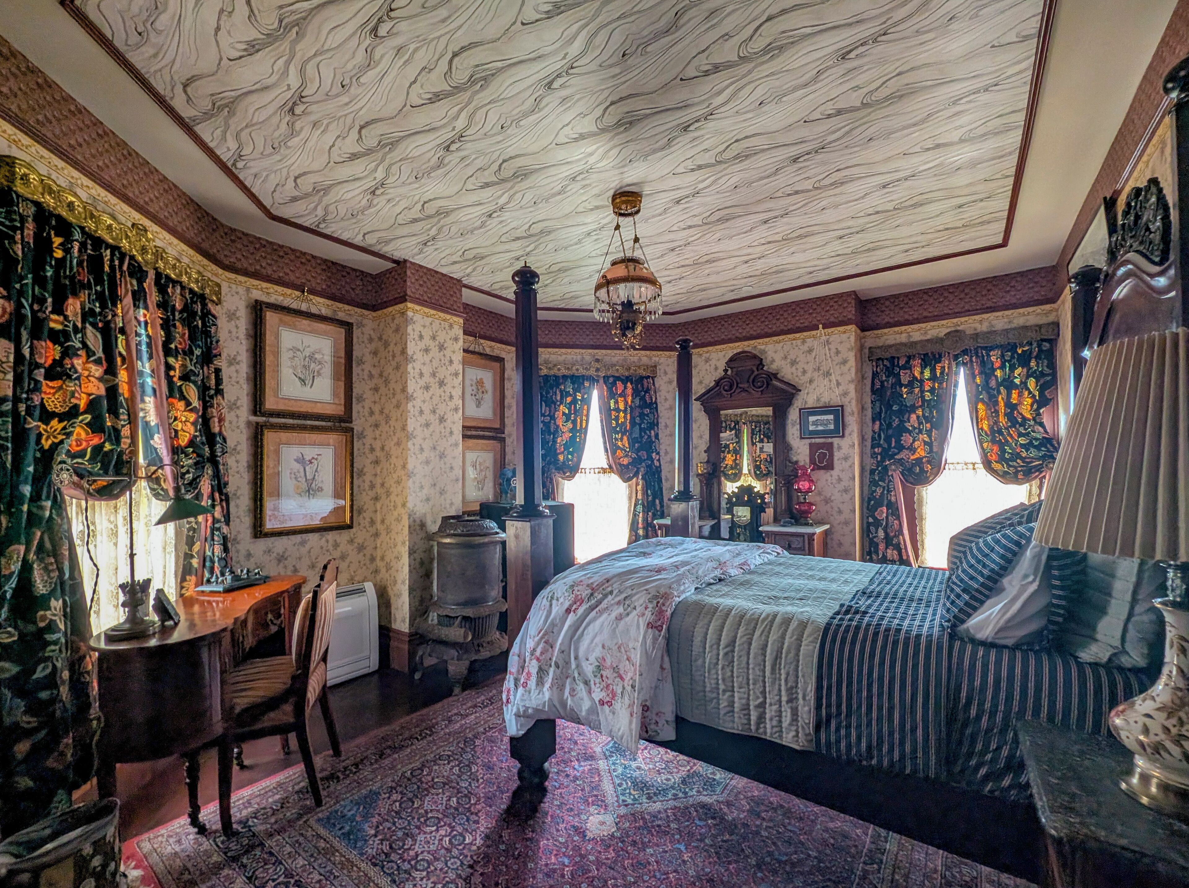 Una habitación con una cama grande, una silla de madera, un escritorio y una alfombra estampada.