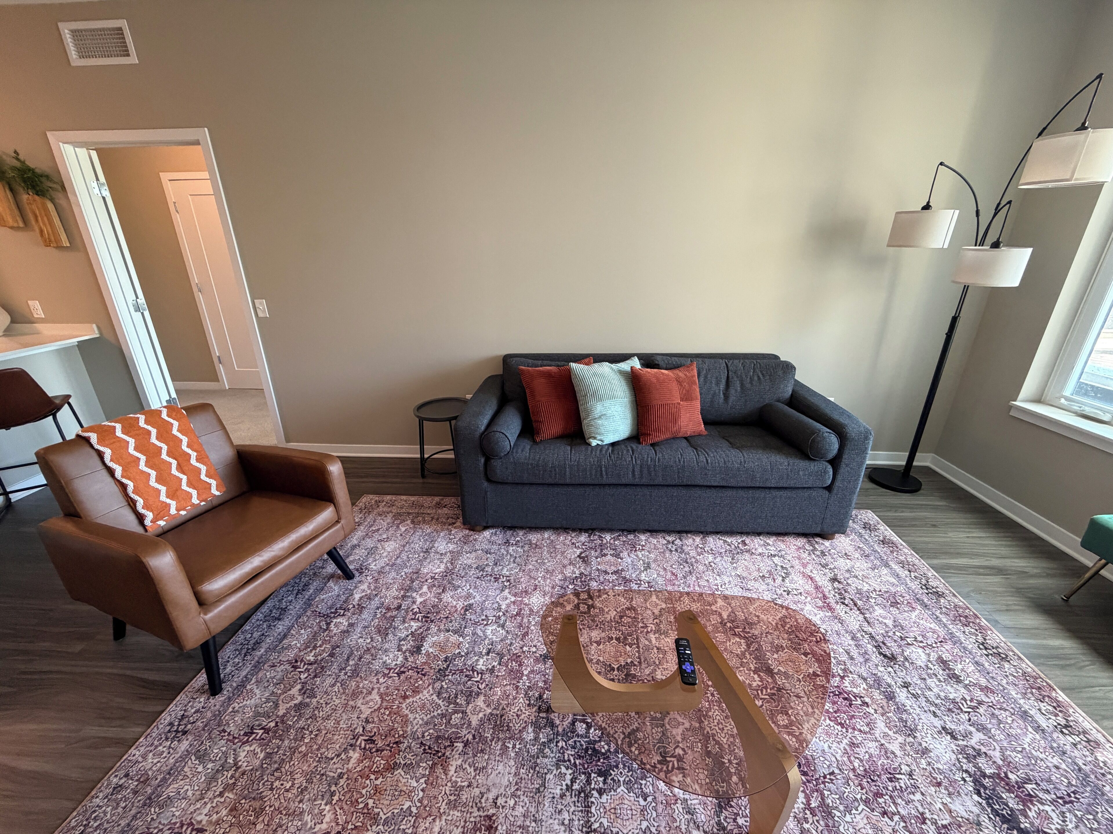 A living room with a brown leather chair, a grey sofa with colorful cushions, a patterned rug, and a tall floor lamp.