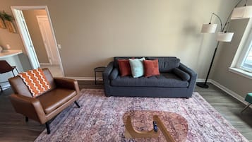 A living room with a brown leather chair, a grey sofa with colorful cushions, a patterned rug, and a tall floor lamp.