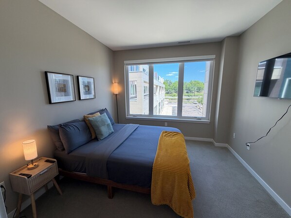 A bedroom with a bed, bedside lamps, a window with a view, and a flat-screen TV mounted on the wall.