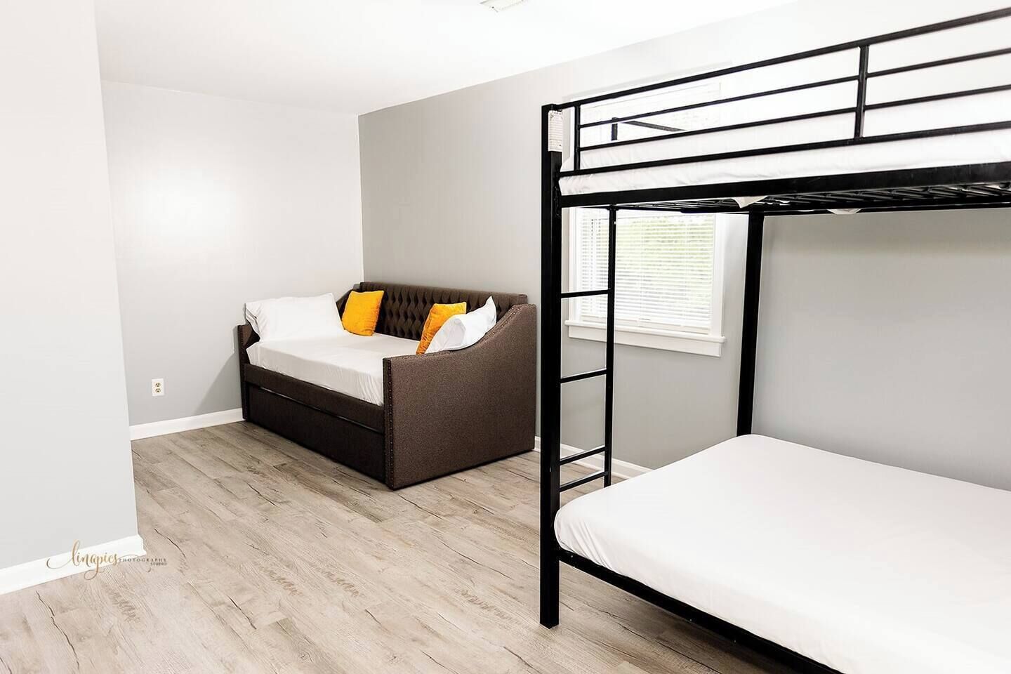 Une pièce avec un lit superposé, un canapé avec des coussins, une fenêtre avec des stores et un plancher en bois.