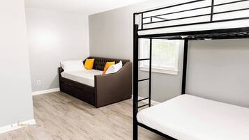 Une pièce avec un lit superposé, un canapé avec des coussins, une fenêtre avec des stores et un plancher en bois.
