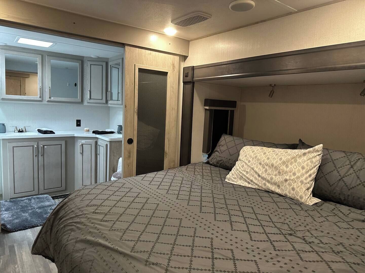 A compact RV bedroom with a bed, pillows, a small kitchenette, and a door leading to another room.