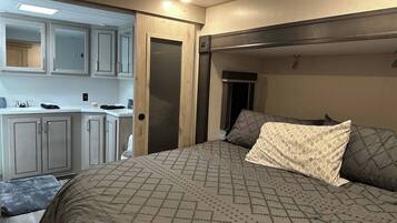 A compact RV bedroom with a bed, pillows, a small kitchenette, and a door leading to another room.