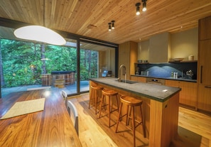 A modern kitchen with wooden cabinetry, a central island with stools, a large window, and a view of a forest.