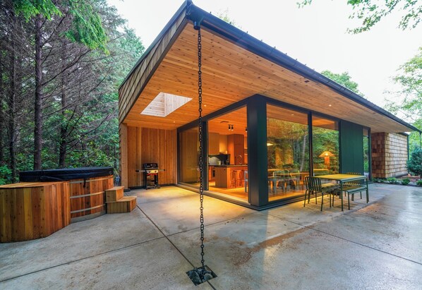 A modern wooden house with a large glass door entrance, a patio with outdoor furniture, and a hot tub.