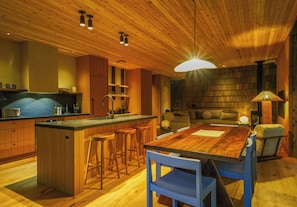 A modern kitchen with wooden cabinetry, a central island with stools, a dining table with chairs, and a cozy seating area with a sofa and cushions.