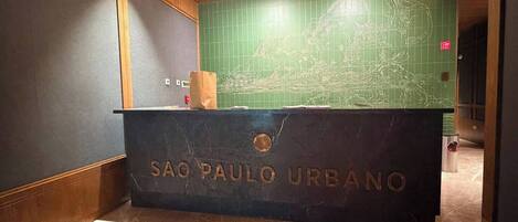 Vastaanottotiski, johon on kaiverrettu teksti »SAO PAULO URBANO», vihreillä laatoilla päällystetty seinä, jossa on kartta, ja puinen katto.