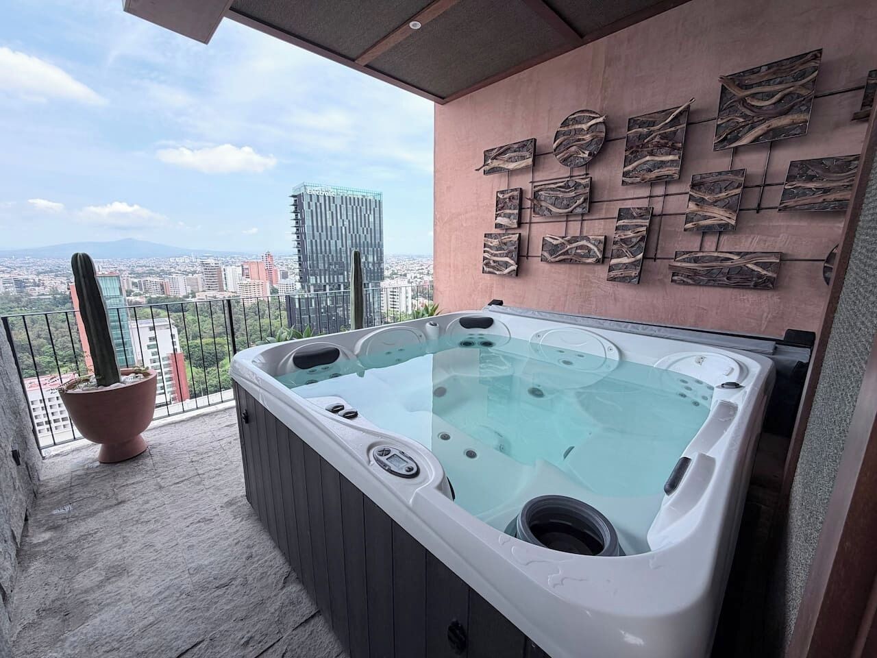 Jacuzzi en un balcón con vistas a la ciudad, una planta en maceta y una pared con arte abstracto.