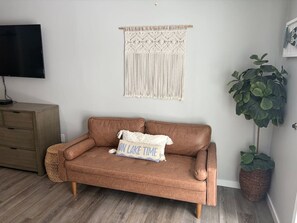 Ein Wohnzimmer mit einem braunen Ledersofa, einem Wandbehang aus Makramee, einem Flachbildfernseher, einem Holzschrank und einem Blumentopf.