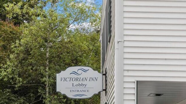 A Victorian Inn entrance sign, a white building with a green porch, and a landscaped garden with various plants. - Victorian Inn #226 | 2 Queen Beds (Elkhart Lake)