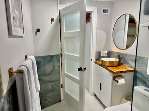 Une salle de bain avec un lavabo blanc, un miroir rond, un porte-serviettes et une porte donnant sur une autre pièce.
