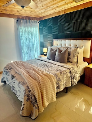 A bedroom with a bed, a nightstand, a lamp, a window with curtains, and a wooden ceiling. - Wood & White (Hialeah)