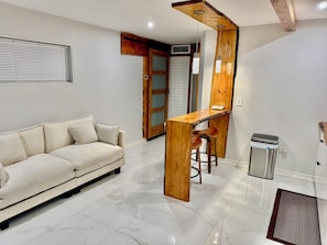 A modern living room with a sofa, wooden bar, stools, and a trash bin. - Wood & White (Hialeah)