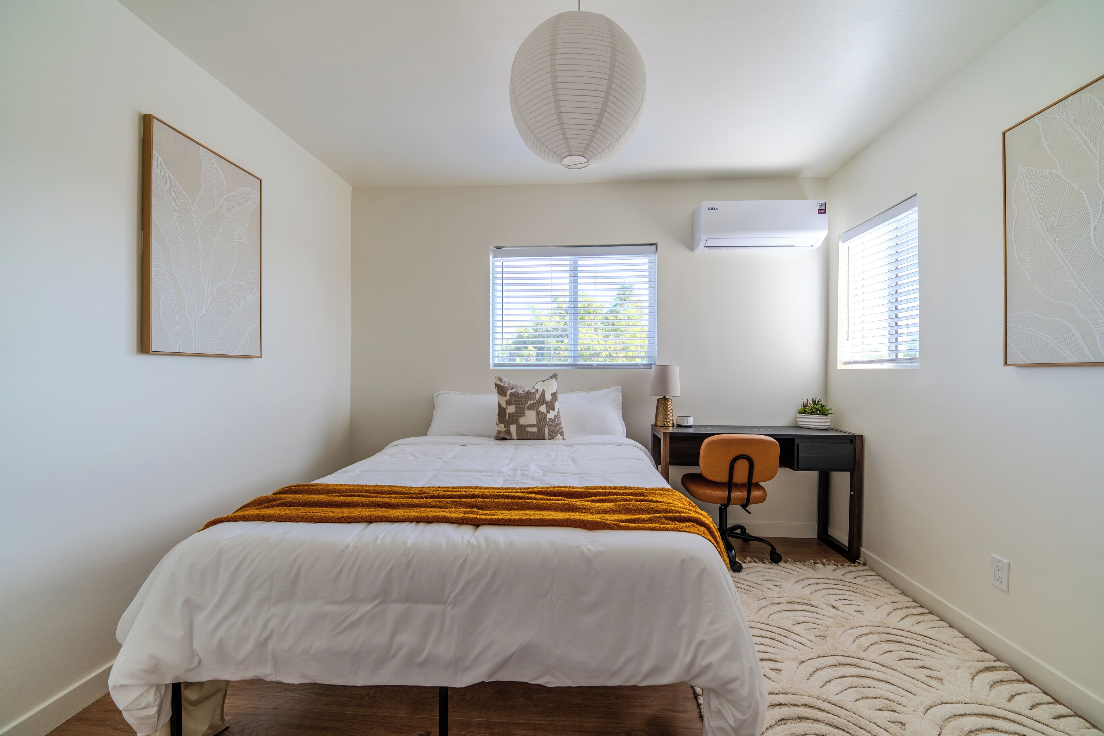 A bedroom with a bed, a desk, and a chair. There is a window with blinds, an air conditioning unit, and a hanging light fixture.