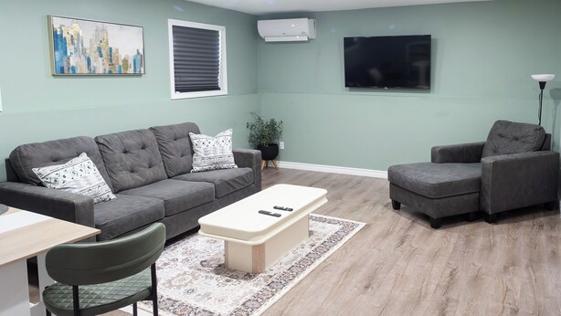 A modern living room with a grey sofa, a matching armchair, a coffee table, and a flat-screen TV mounted on the wall.