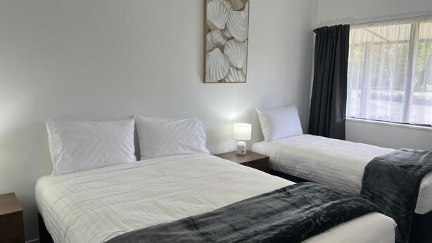 A hotel room with two beds, a nightstand with a lamp, a wall-mounted artwork, and a window with curtains. - Magnolia Court Motel (Ōpōtiki)