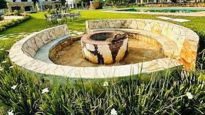 Un foyer circulaire en pierre, entouré de gazon et de fleurs, avec une piscine et des bâtiments en arrière-plan.