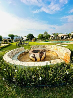 A circular stone fire pit surrounded by grass and flowers, with a pool and buildings in the background.