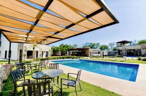 A swimming pool with a covered patio area, outdoor seating, and a modern building in the background.