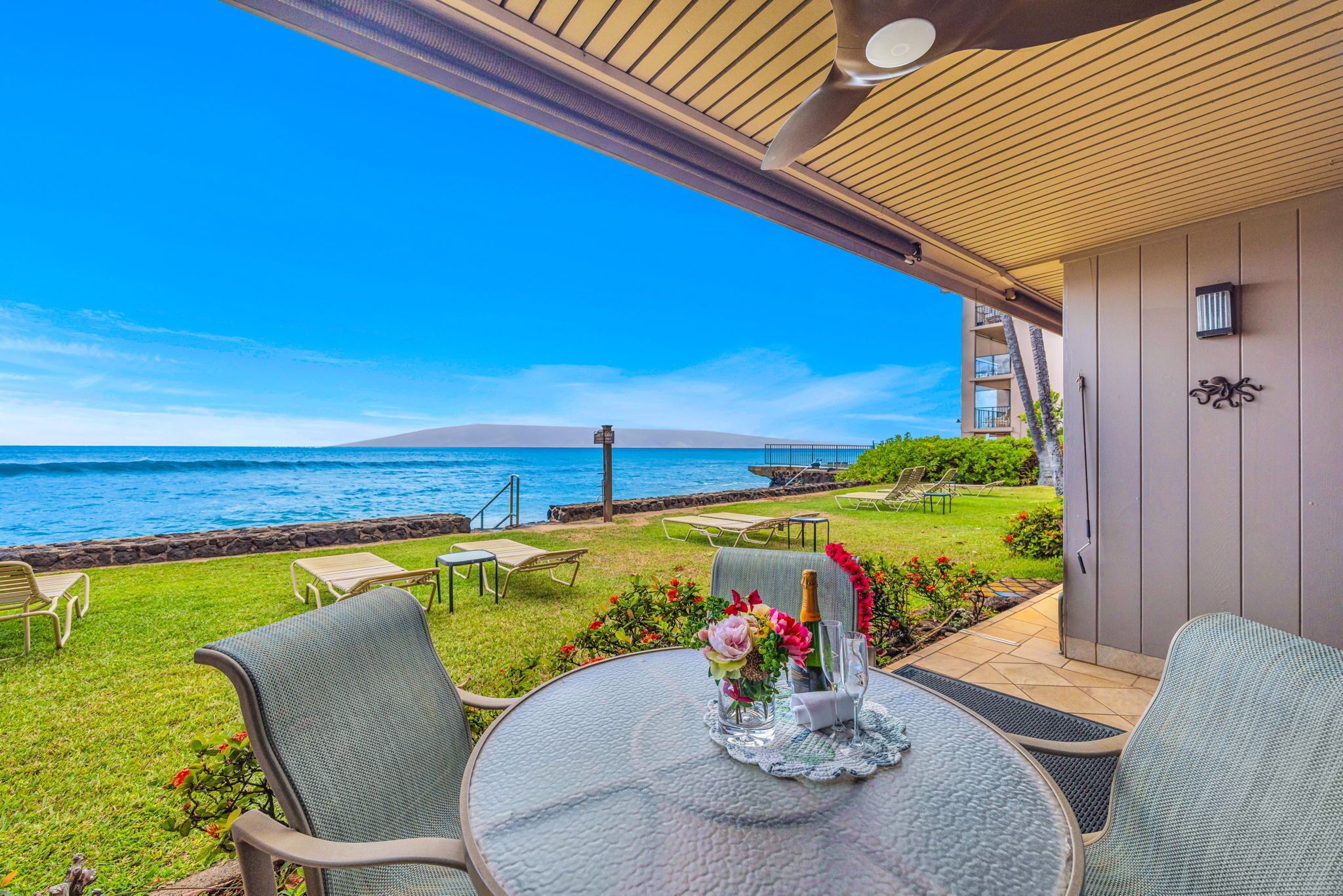 An outdoor patio with a table set for two, overlooking a beach with a clear view of the ocean and a distant island.
