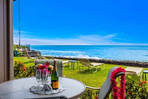 A table with a bottle of champagne, glasses, and a decorative item, overlooking a beach with lounge chairs and a clear view of the ocean. - Beachfront at The Pikake, ocean views (Lahaina)