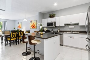 A modern kitchen with white cabinets, a central island, bar stools, and decorative artwork. - Family-Friendly 3BR Townhouse with Outdoor Patio & Fire Pit 🌴 (Cape Canaveral)
