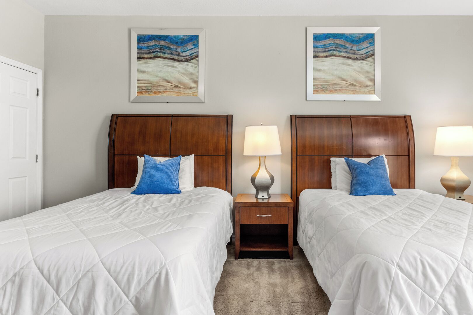 A hotel room with two beds, wooden headboards, white bedding, blue pillows, bedside tables with lamps, and two framed artworks on the wall.