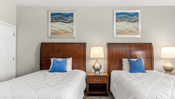 Ein Hotelzimmer mit zwei Betten, Holz-Kopfteilen, weißer Bettwäsche, blauen Kissen, Nachttischen mit Lampen und zwei gerahmten Kunstwerken an der Wand.
