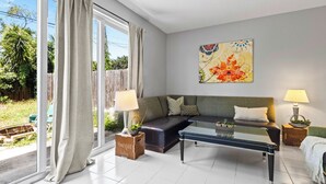 Ein Wohnzimmer mit einer Couch, einem Couchtisch und zwei Lampen. Ein Fenster mit Vorhängen, ein Bild an der Wand und Blick ins Grüne sind zu sehen.