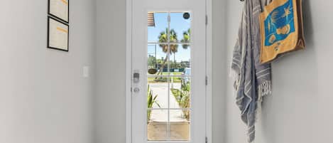 A hallway with a white door, a view of palm trees and a parking lot, and a wall with a towel rack and a framed certificate.